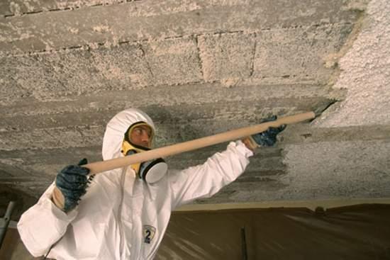 Asbestos abatement worker in a protective suit and respirator scraping a ceiling